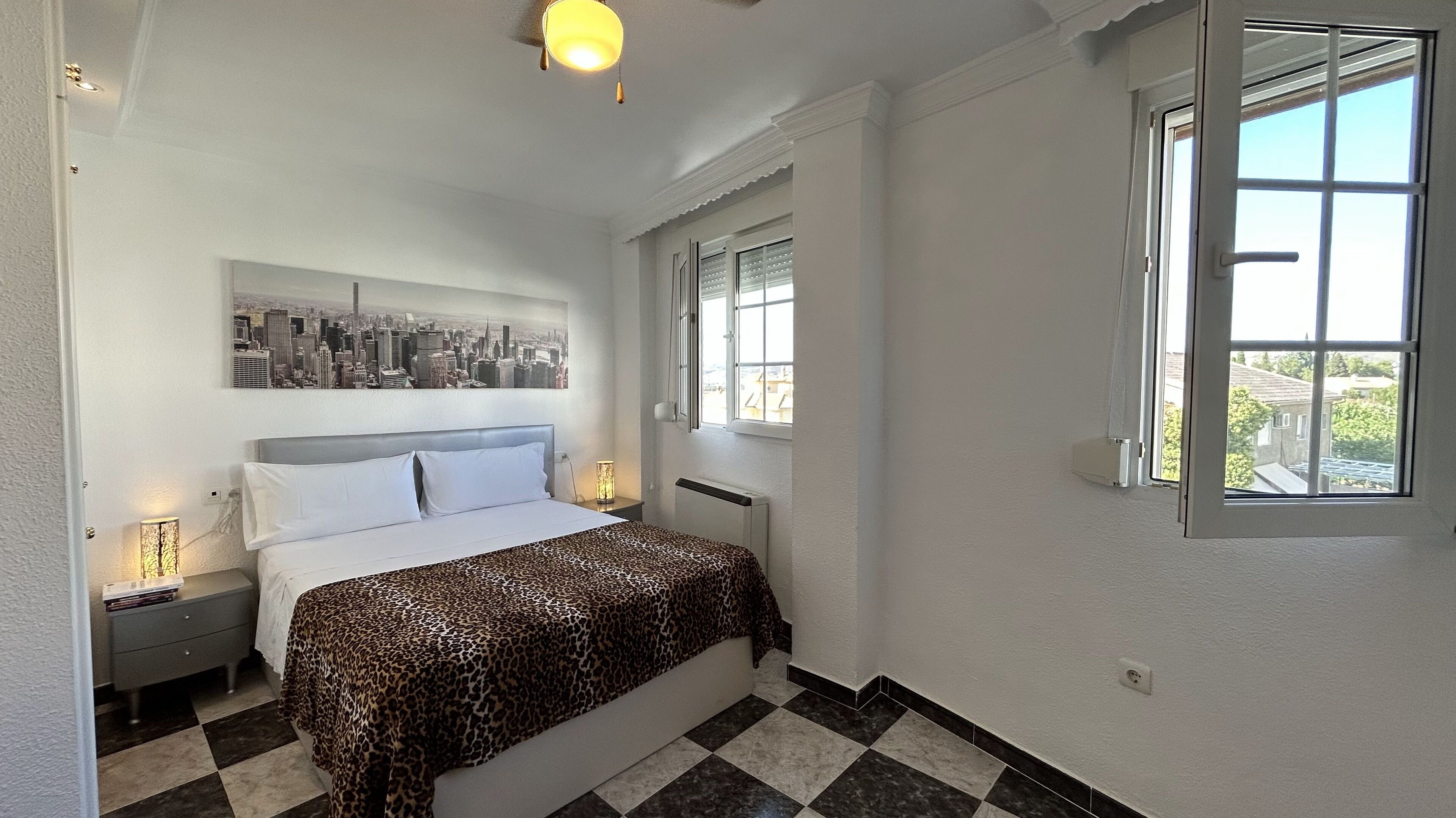 Un dormitorio con una cama, una obra de arte con un paisaje urbano, un radiador, una ventana y un suelo a cuadros.