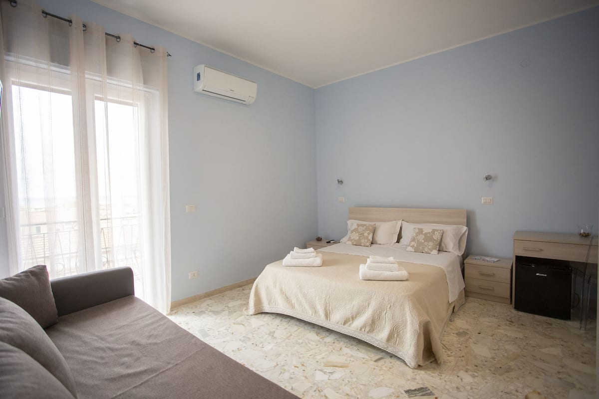 A bedroom with a bed, a bedside table, a small cabinet, and a window with curtains.