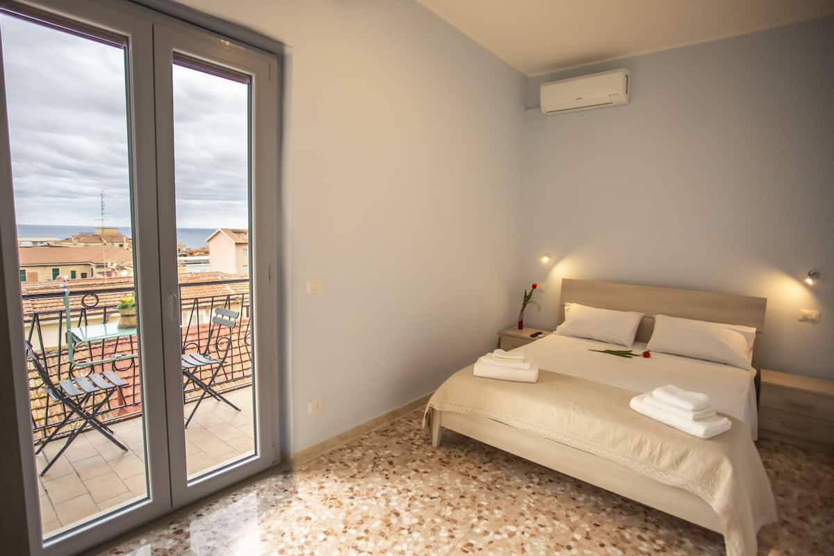 A bedroom with a bed, a nightstand, and a window overlooking a balcony with chairs and a table.