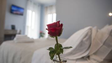 A single red rose in a clear glass vase on a bed in a hotel room.