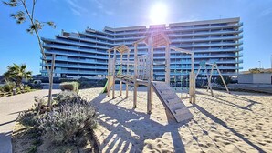 Un’area giochi sulla spiaggia con una struttura in legno e una zona sabbiosa, di fronte a un moderno hotel a più piani.