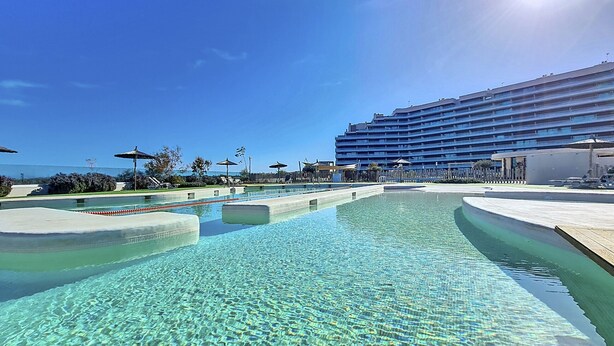 Una grande piscina con acqua cristallina, circondata da un edificio di un hotel e zone relax.