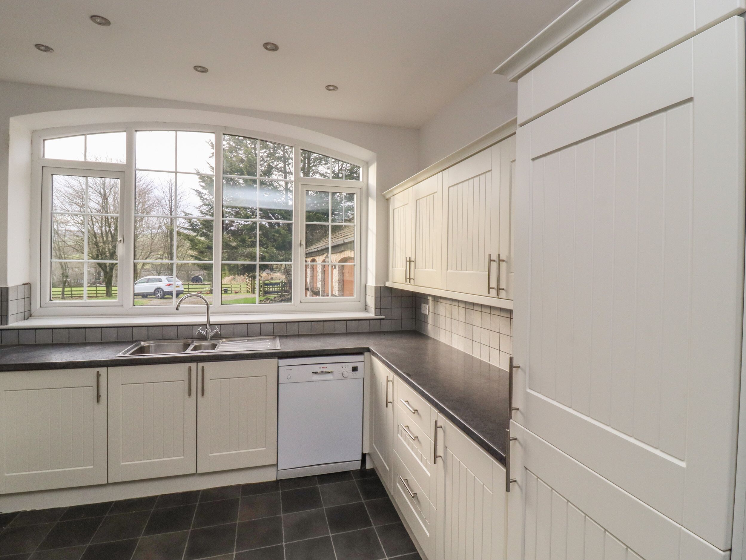 A kitchen with white cabinets, a dishwasher, and a window with a view of a car and trees.