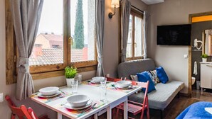 A cozy room with a dining table set for two, a sofa with colorful cushions, and a TV mounted on the wall. - Apartamento en Pleno Centro de Navacerrada (Navacerrada)