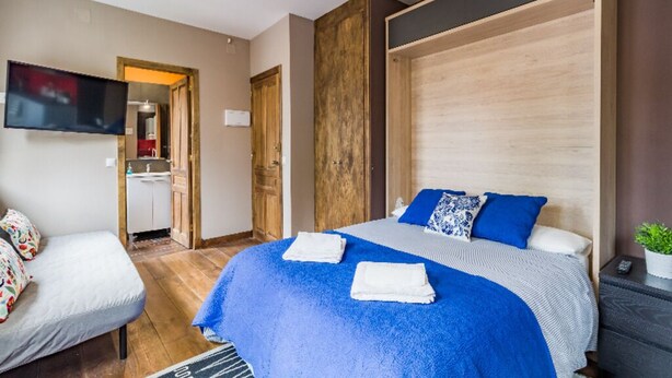 A modern hotel room with a large bed, a TV mounted on the wall, a small kitchenette, and a wooden accent wall. - Apartamento en Pleno Centro de Navacerrada (Navacerrada)