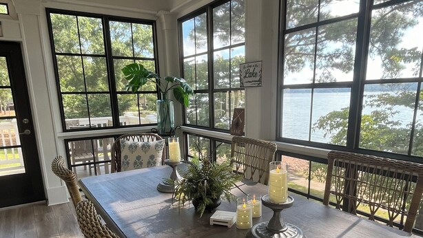 A dining area with a table set for four, surrounded by wicker chairs, with a view of a lake through large windows. - Great Waters gem! Spacious Waterfront Cottage! Family Friendly Getaway! (Eatonton)