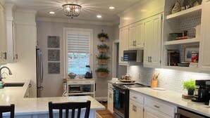 A modern kitchen with white cabinetry, a central island with two chairs, stainless steel appliances, and a chandelier. - Great Waters gem! Spacious Waterfront Cottage! Family Friendly Getaway! (Eatonton)