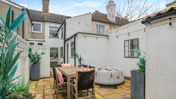 A courtyard with a wooden table and chairs, a white sofa, potted plants, and a white wall.
