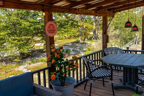 Een houten terras met een tafel en stoelen, omringd door een reling, met uitzicht op een natuurlandschap met bomen en een beek.