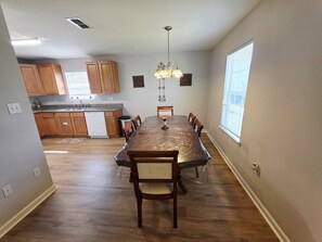 A dining area with a long table, wooden chairs, a chandelier, and a kitchen in the background.
