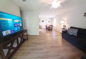 A living room with a flat-screen TV on a wooden stand, a dark-colored sofa, and a dining area with a chandelier.