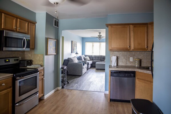 A kitchen with wooden cabinets, a microwave, an oven, and a dishwasher. There is a living room area with a sofa and a ceiling fan.