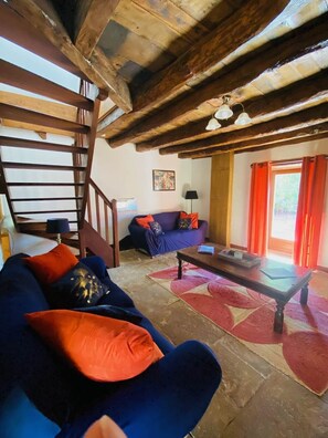 Un interior rústico con una escalera de madera, un sofá azul con cojines naranja, una mesa de centro de madera y una alfombra roja con estampado.