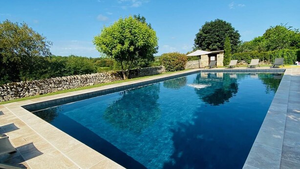 Una piscina rectangular con borde de piedra, rodeada por un muro de piedra y árboles.