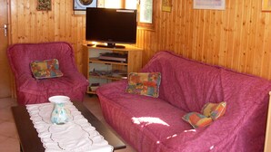 A cozy room with a wooden interior, featuring a pink sofa, a television, and a small table with a vase. - Location Vacances des Quatre Vents (GIRMONT VAL D AJOL)