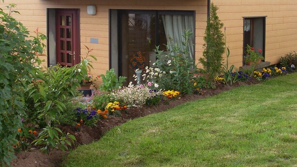 A two-story house with a balcony, a front door, and a garden with various plants and flowers. - Location Vacances des Quatre Vents (GIRMONT VAL D AJOL)