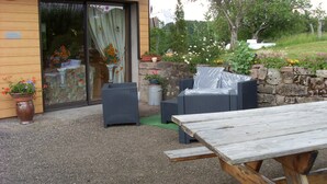 An outdoor patio with wicker furniture, a wooden table, and potted plants. - Location Vacances des Quatre Vents (GIRMONT VAL D AJOL)