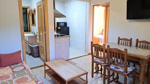 A compact living space with a kitchenette, a dining area with wooden chairs, a small table, and a sofa. - Apartamento Rural en Plena Ribeira Sacra (Cerreda)