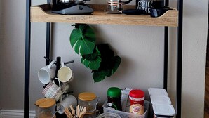 A coffee station with a coffee maker, grinder, and various coffee-related items on a wooden shelf against a wall with a green leafy backdrop and a neon sign that reads "BUT FIRST Coffee." - Quiet Lakeside home w/bbq close to Daytona & Sanford  (Deltona)