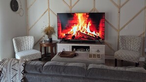 A living room with a TV displaying a fireplace, two patterned chairs, a grey sofa, and a small wooden side table with a vase. - Quiet Lakeside home w/bbq close to Daytona & Sanford  (Deltona)