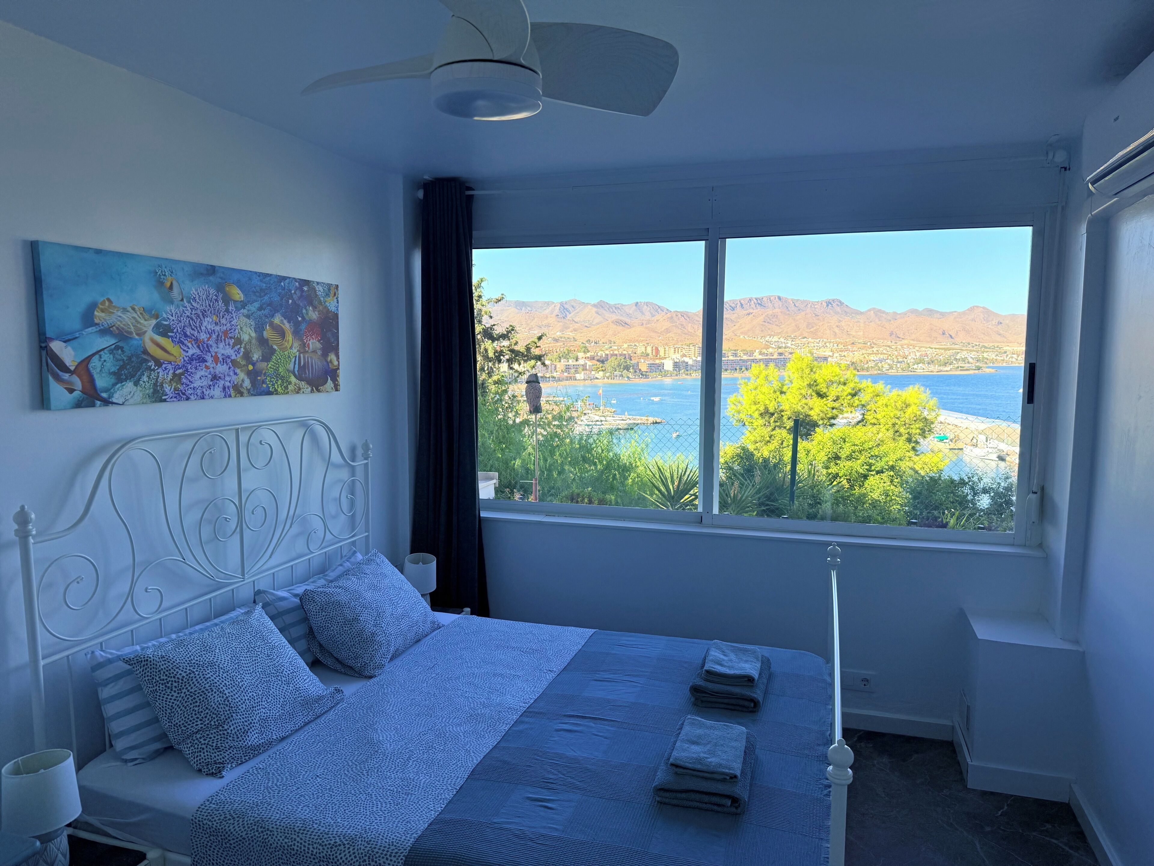 Ein Schlafzimmer mit einem Bett, Deckenventilator, einem Gemälde und Blick auf ein Gewässer und Berge.