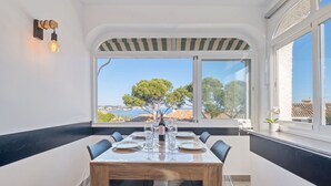 Un coin repas avec une table dressée pour quatre personnes, de grandes fenêtres offrant une vue sur la mer et les arbres, et un luminaire moderne fixé au mur.