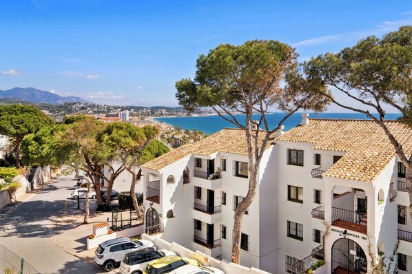 Un bâtiment situé sur la côte, doté de nombreux balcons, une rue pavée, des voitures garées et une vue sur la mer et les montagnes lointaines.