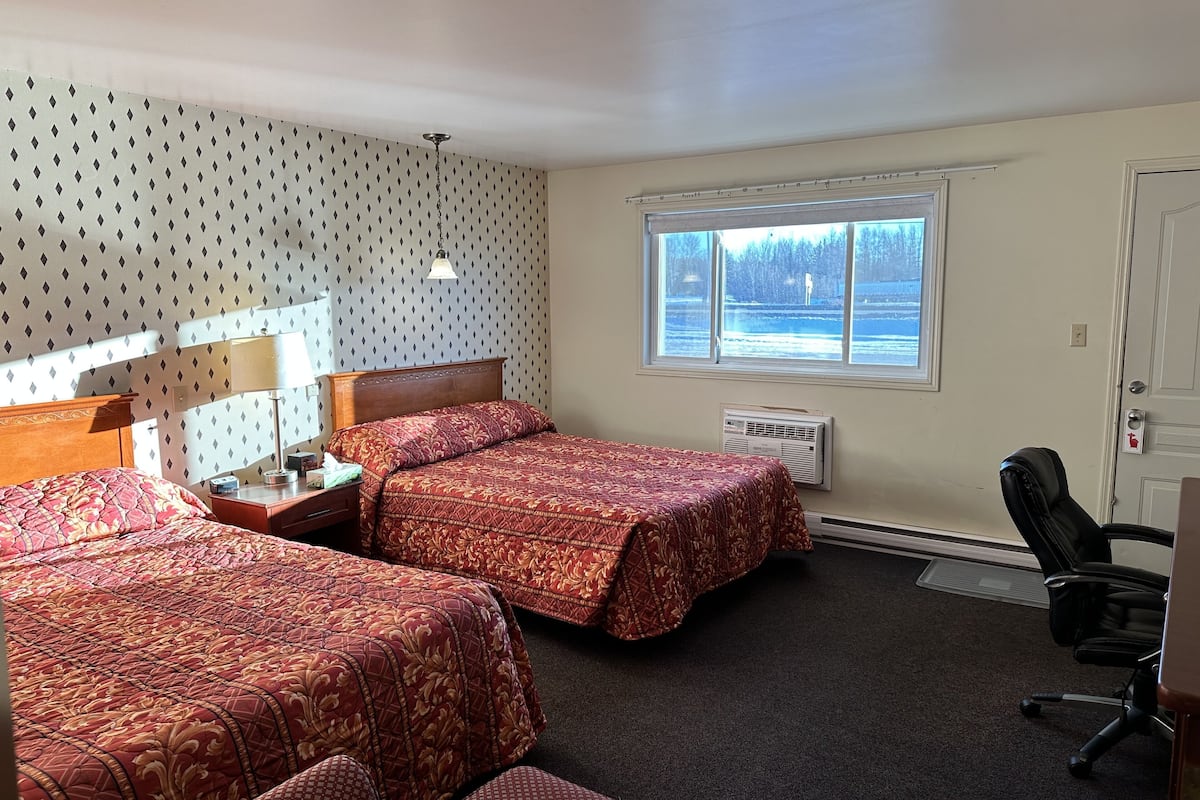 A hotel room with two beds, a desk, a chair, a window with a view of a snowy landscape, and a radiator.