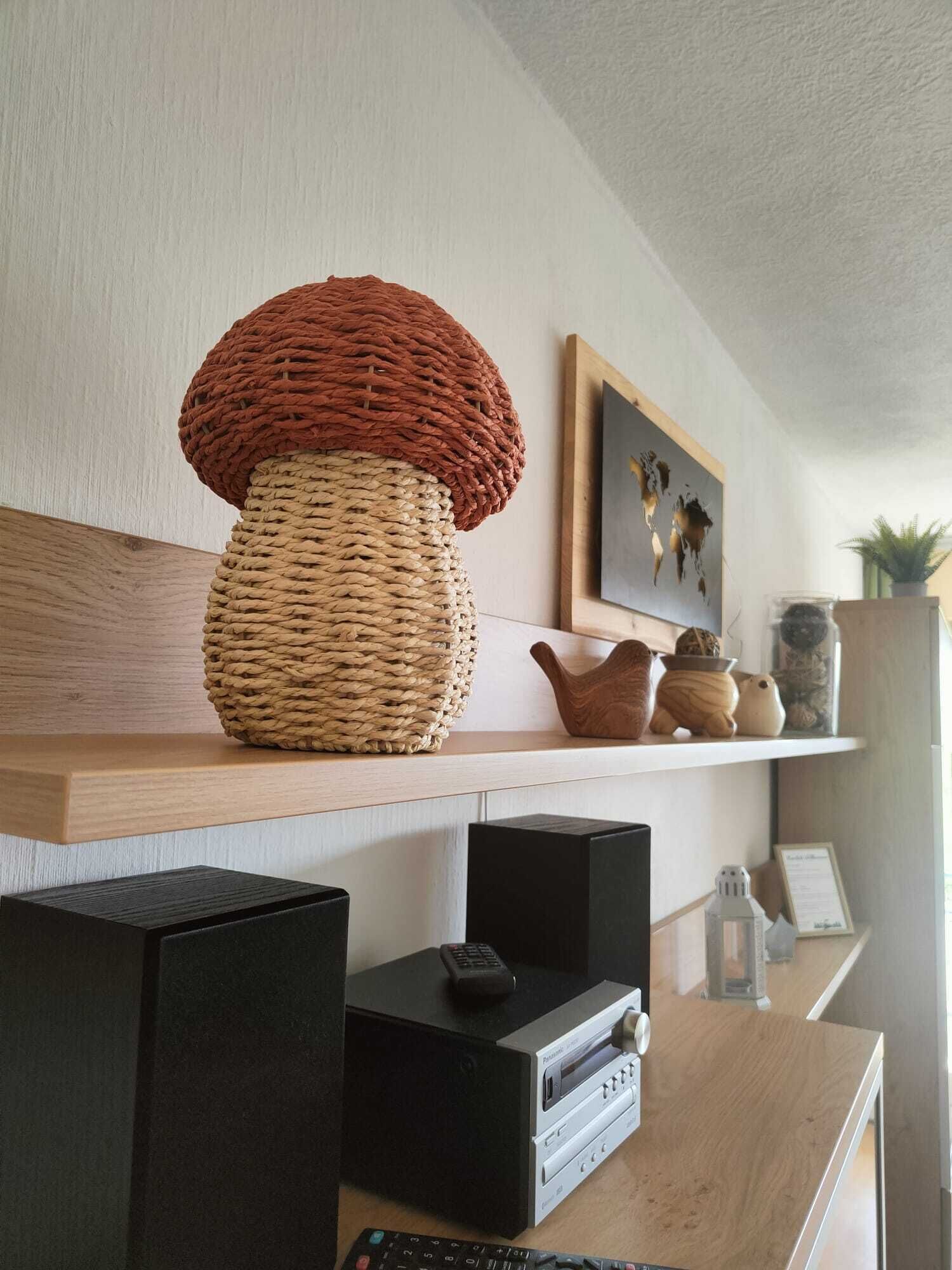Een werkplek aan huis met een grote geweven lamp, een ingelijst kunstwerk en een plank met decoratieve objecten.
