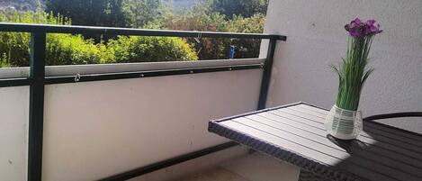 A balcony with a table and chairs, a vase with flowers, and a view of greenery and hills.
