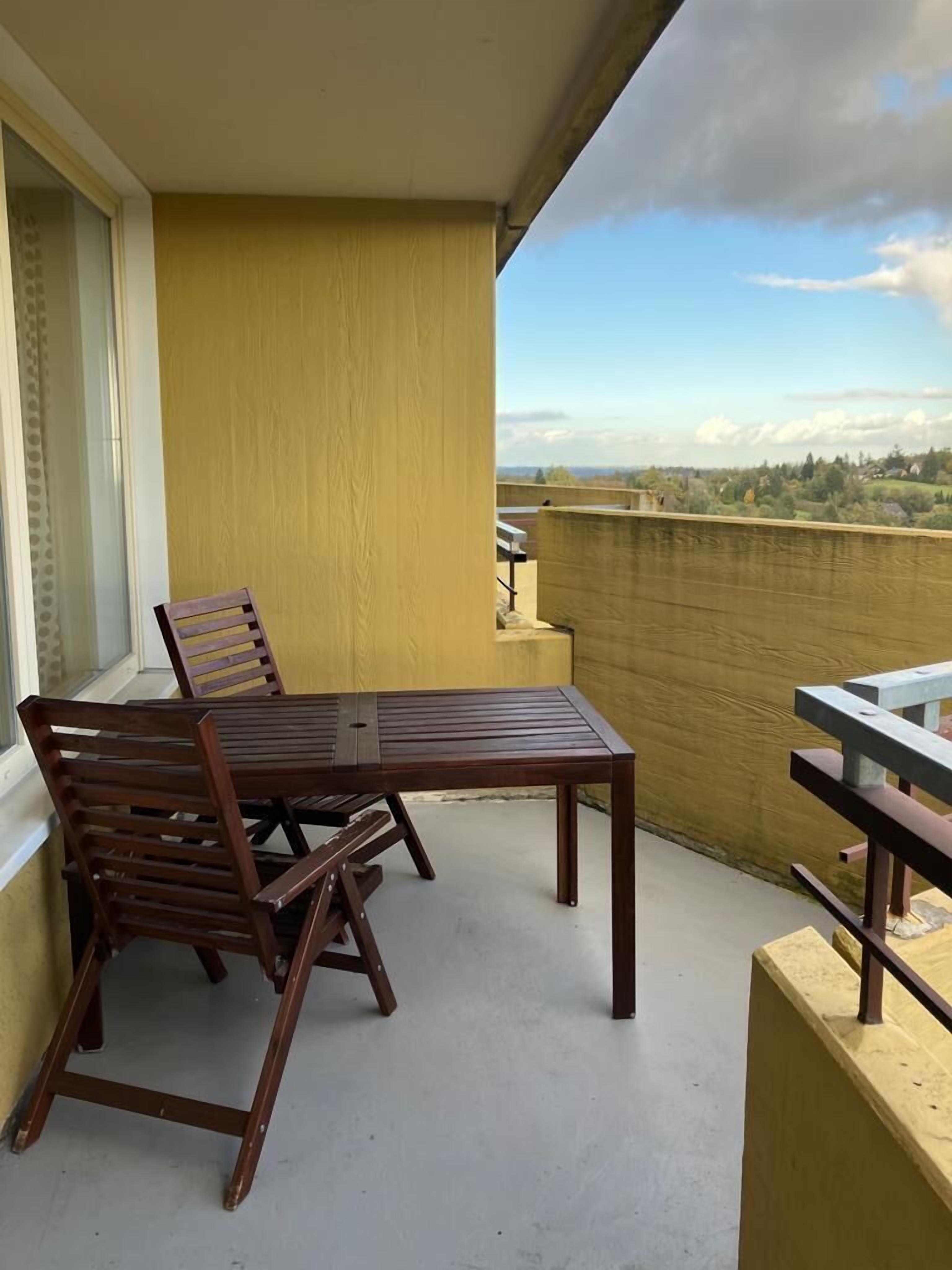 Un balcón con una mesa y sillas de madera, vista a un paisaje lejano y una pared amarilla.