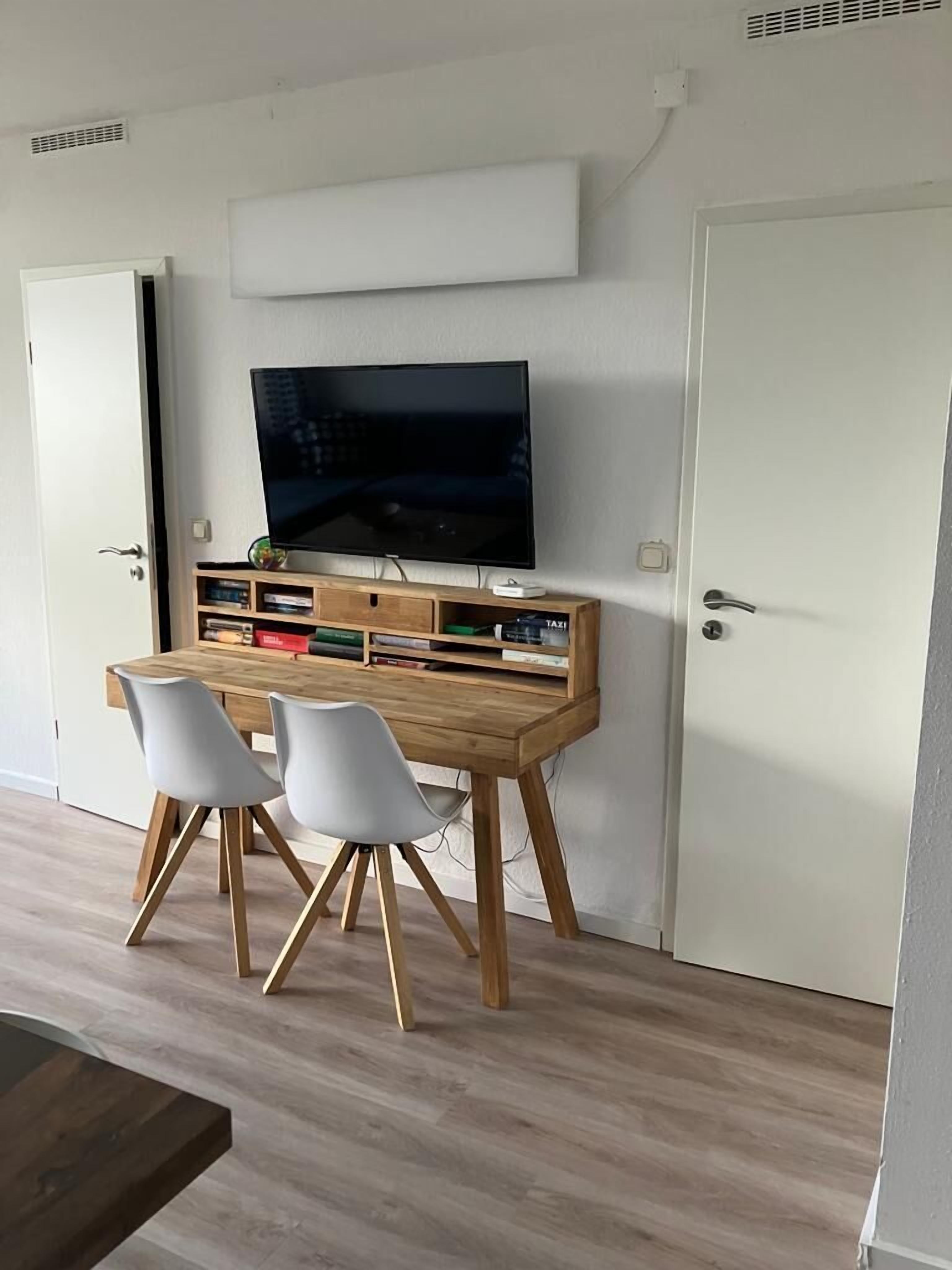 Une pièce moderne comprenant un bureau en bois, deux chaises blanches, un téléviseur à écran plat et une bibliothèque.