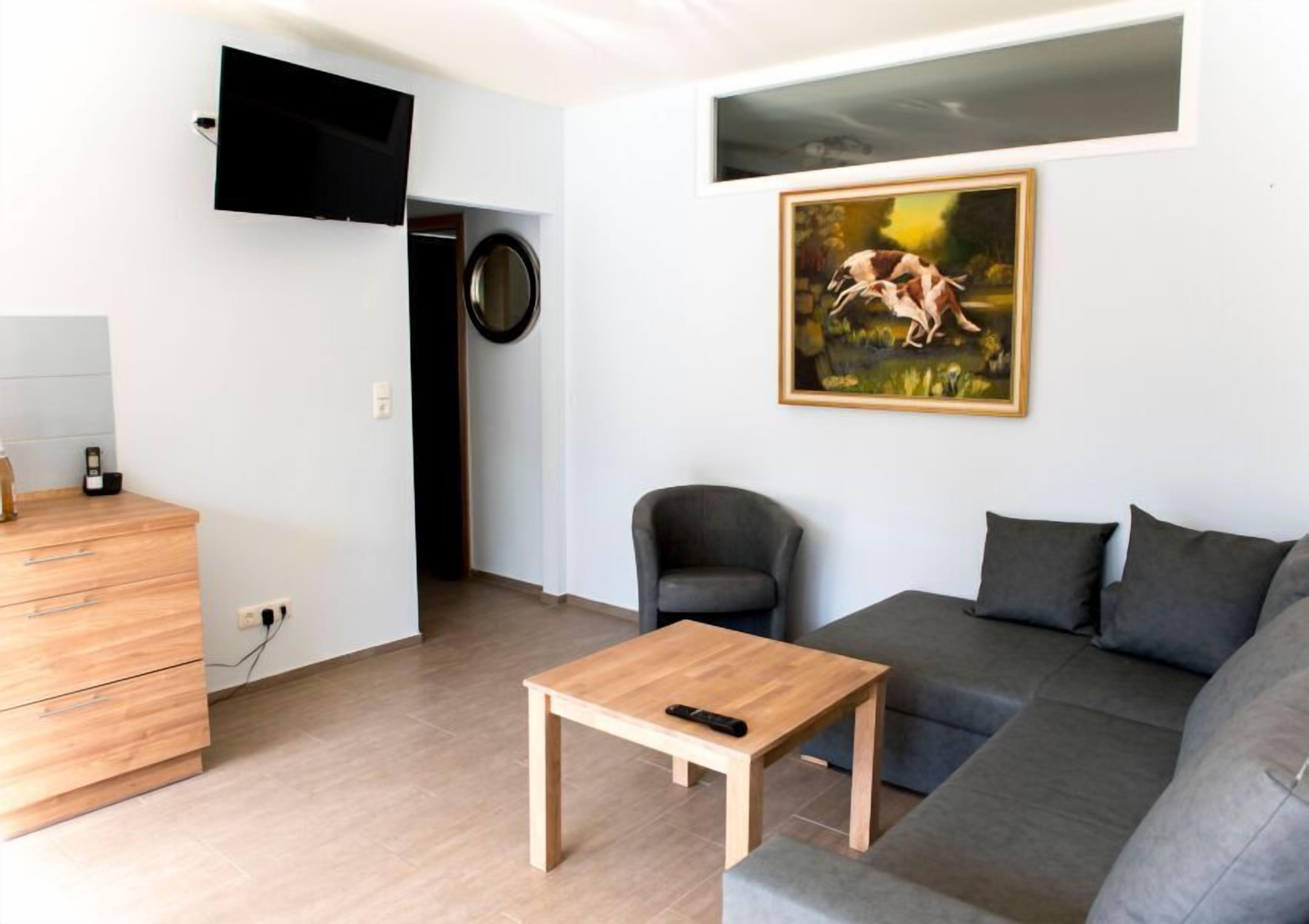 A modern living room with a grey sofa, a wooden coffee table, a black armchair, a TV mounted on the wall, and a painting.