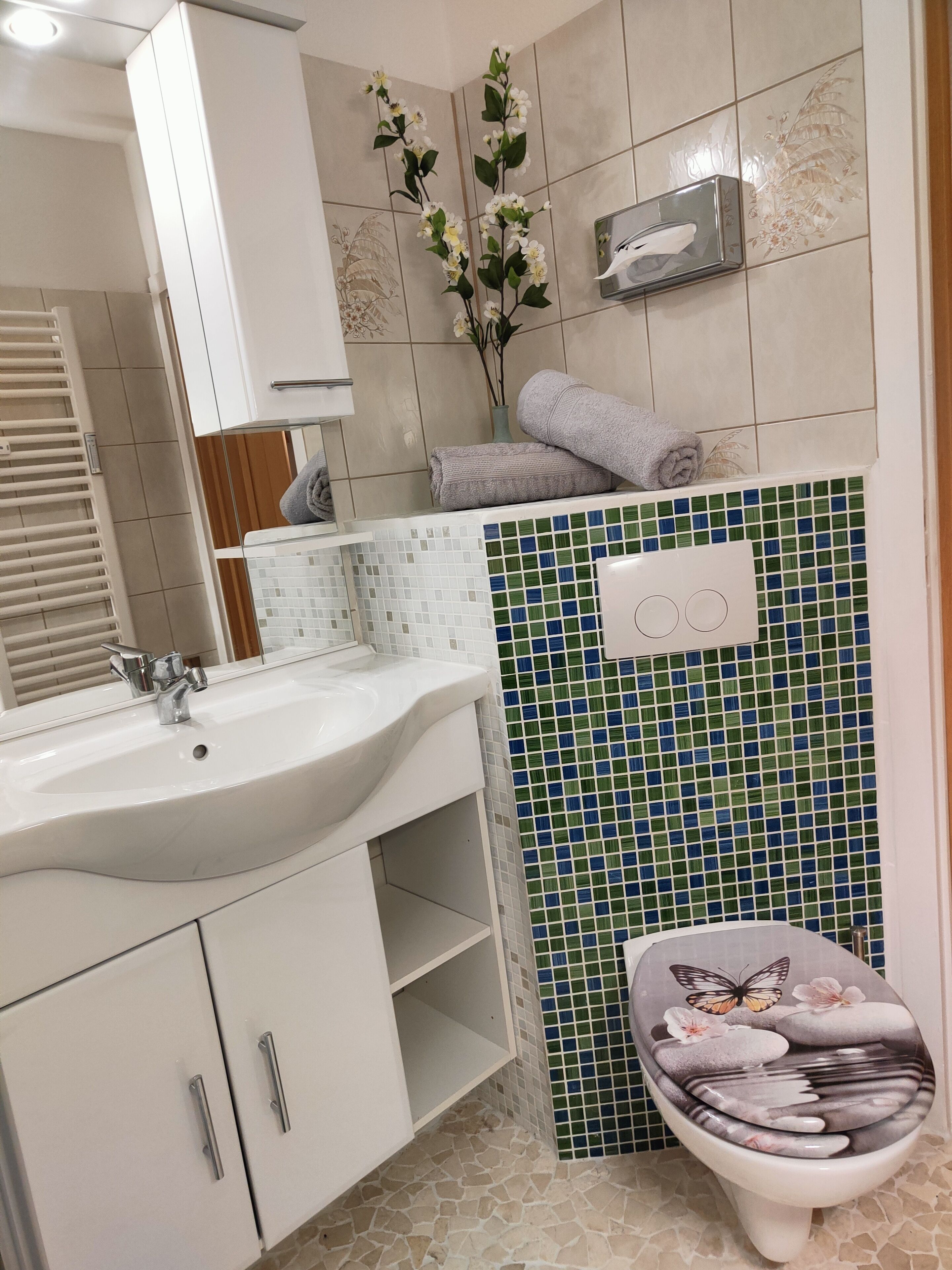 Een badkamer met een witte wastafel, een spiegel, een toilet met een decoratieve zitting en een betegelde muur met groene en blauwe tegels.