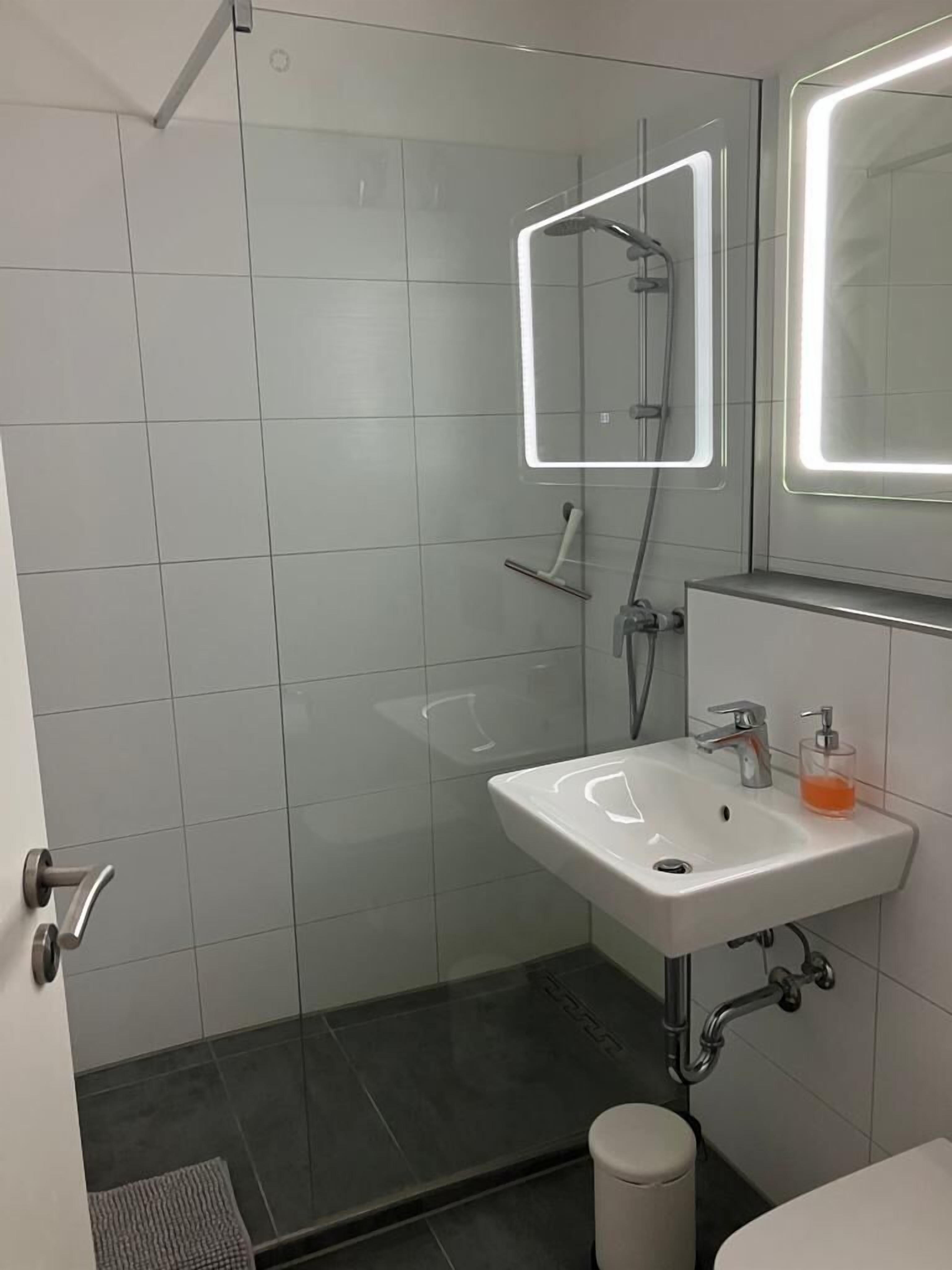 Une salle de bain moderne avec une douche à parois de verre, un lavabo blanc doté d’un distributeur de savon et des toilettes.