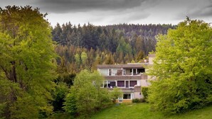 A modern two-story building surrounded by lush greenery and a forested hill in the background. - Apartments Harz Panorama (Braunlage)