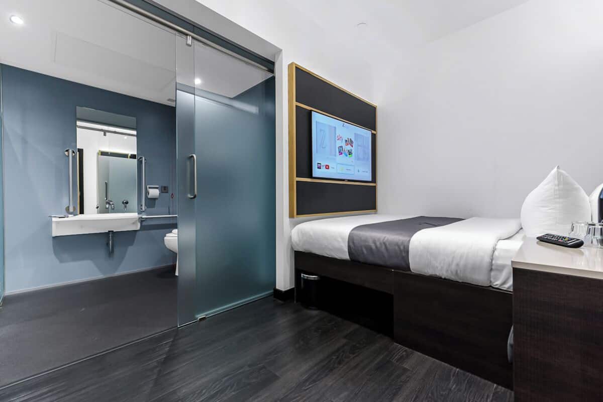 Une chambre d’hôtel moderne dotée d’une paroi en verre séparant la salle de bain, d’un lit avec un panneau de commande et d’une table de chevet avec une télécommande.
