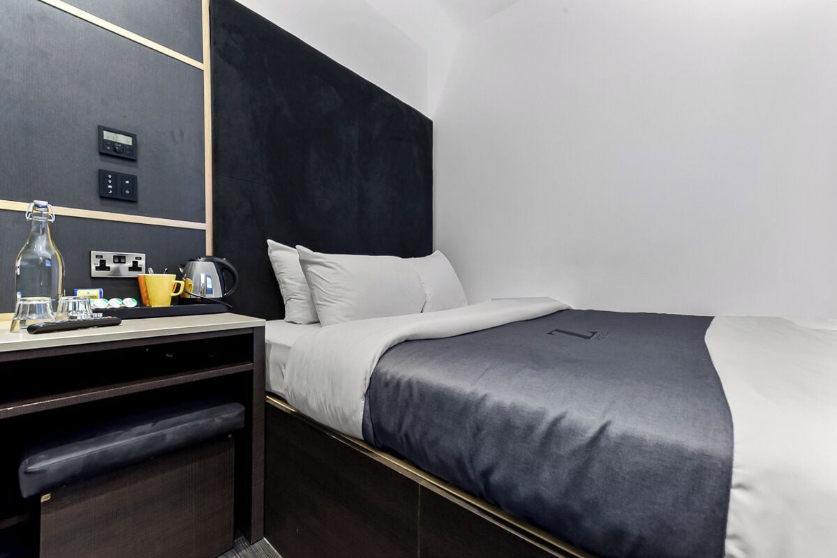 Quarto de hotel moderno com cabeceira escura, cama com roupa de cama branca e cinza, escrivaninha com chaleira e espelho na parede.