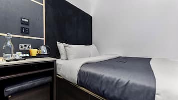 Quarto de hotel moderno com cabeceira escura, cama com roupa de cama branca e cinza, escrivaninha com chaleira e espelho na parede.