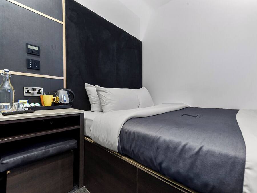 A modern hotel room with a dark headboard, a bed with white and gray bedding, a desk with a kettle, and a mirror on the wall.