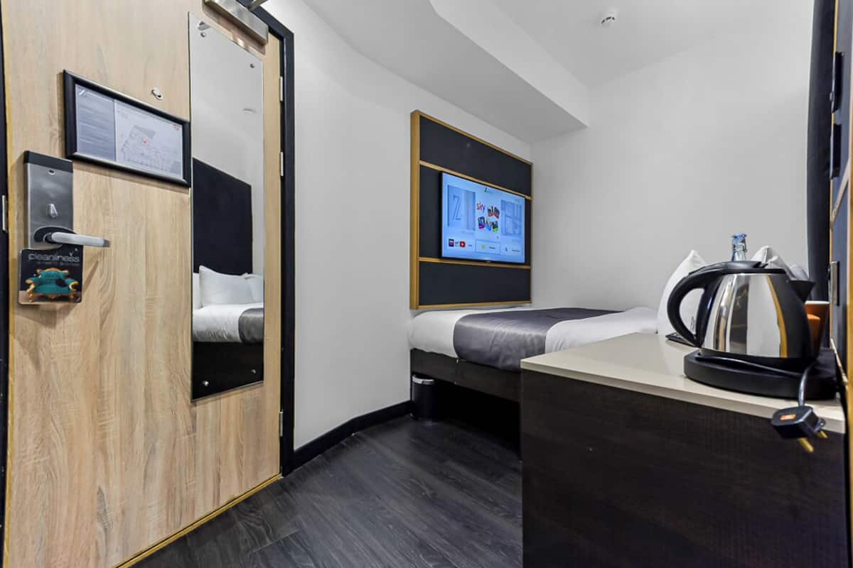Une chambre d’hôtel de taille réduite, équipée d’un lit, d’un bureau, d’une machine à café et d’un écran de télévision affichant une carte.