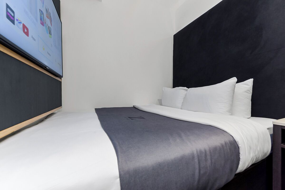 A modern hotel room with a bed, a black headboard, white bedding, and a flat-screen TV mounted on the wall.