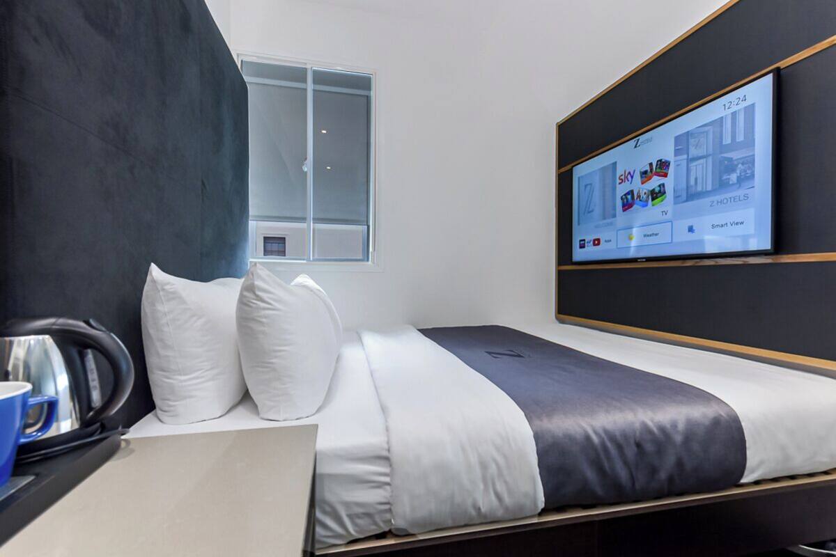 Une chambre d’hôtel moderne équipée d’un lit, d’un téléviseur à écran plat affichant l’application de l’hôtel, d’une fenêtre et d’une bouilloire posée sur la table de chevet.