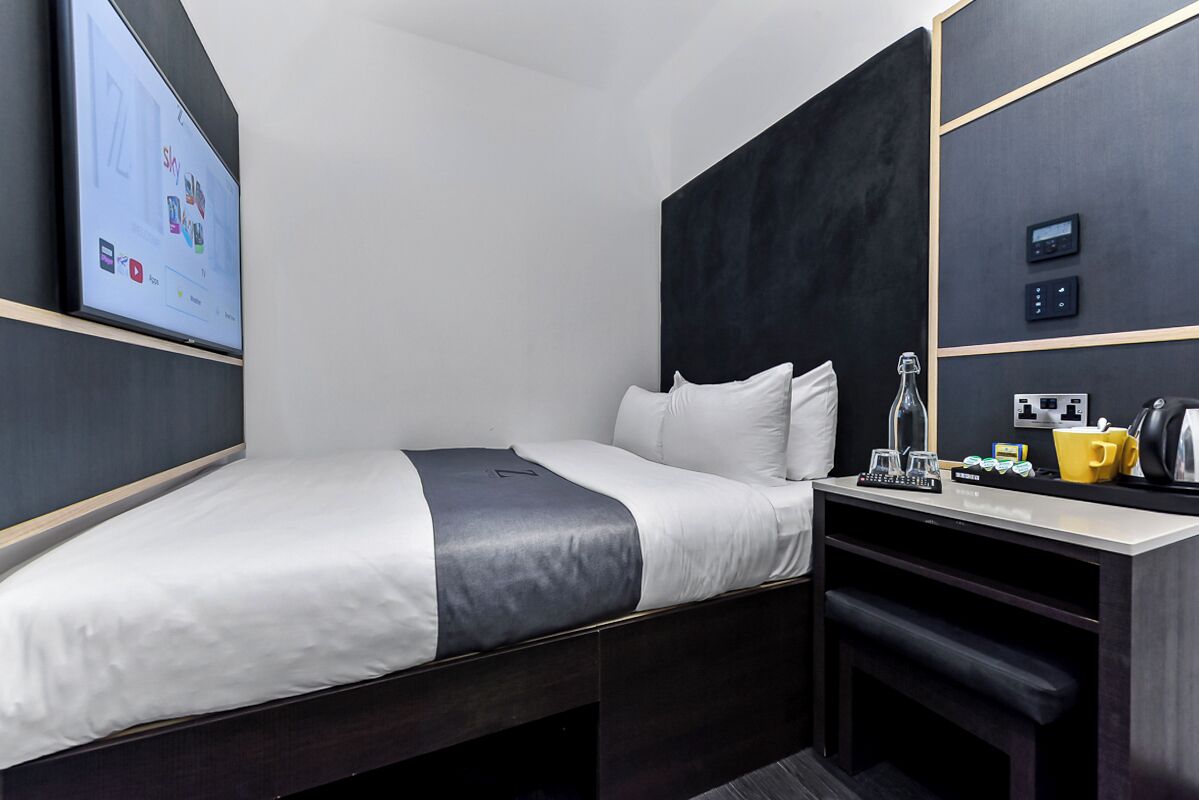 Ein modernes Hotelzimmer mit Bett, Schreibtisch, Kaffeemaschine und digitaler Wanduhr.