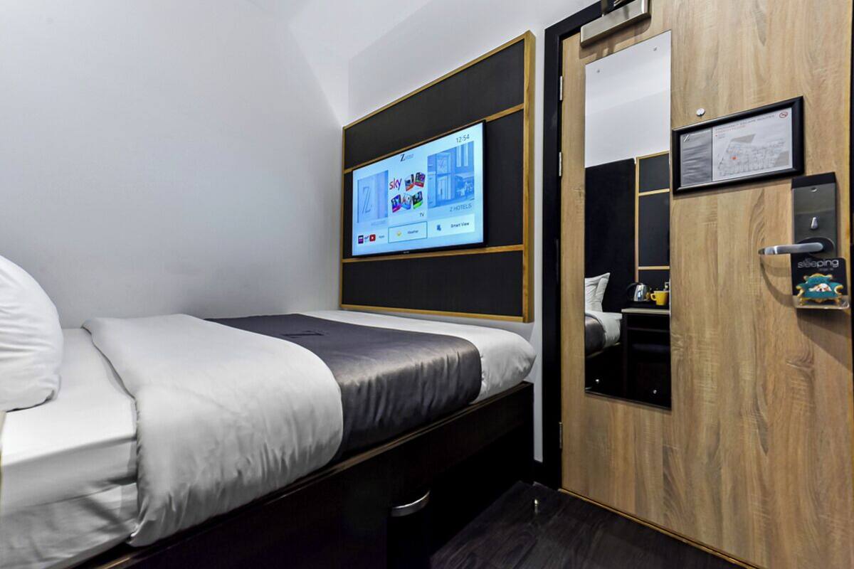 Une chambre d’hôtel moderne équipée d’un lit, d’un téléviseur fixé au mur qui affiche une application de l’hôtel, d’une armoire en bois et d’un petit bureau avec une chaise.