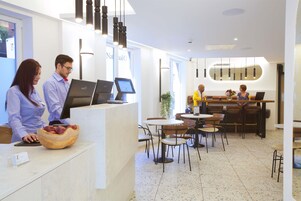 A modern hotel lobby with a reception desk, computers, and a seating area with tables and chairs.
