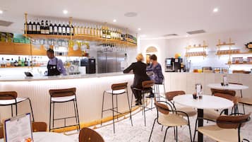 Una zona de bar con mostrador, varias estanterías llenas de botellas y copas. Hay varias sillas y mesas alrededor de la barra.