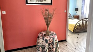 A room with a red wall, a framed picture of boats, a small table with a patterned tablecloth, and a wicker chair.