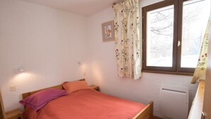 Ein Schlafzimmer mit einem hölzernen Bett, einem Nachttisch, einer Wandlampe, einem Fenster mit floralem Vorhang und einer Heizung.