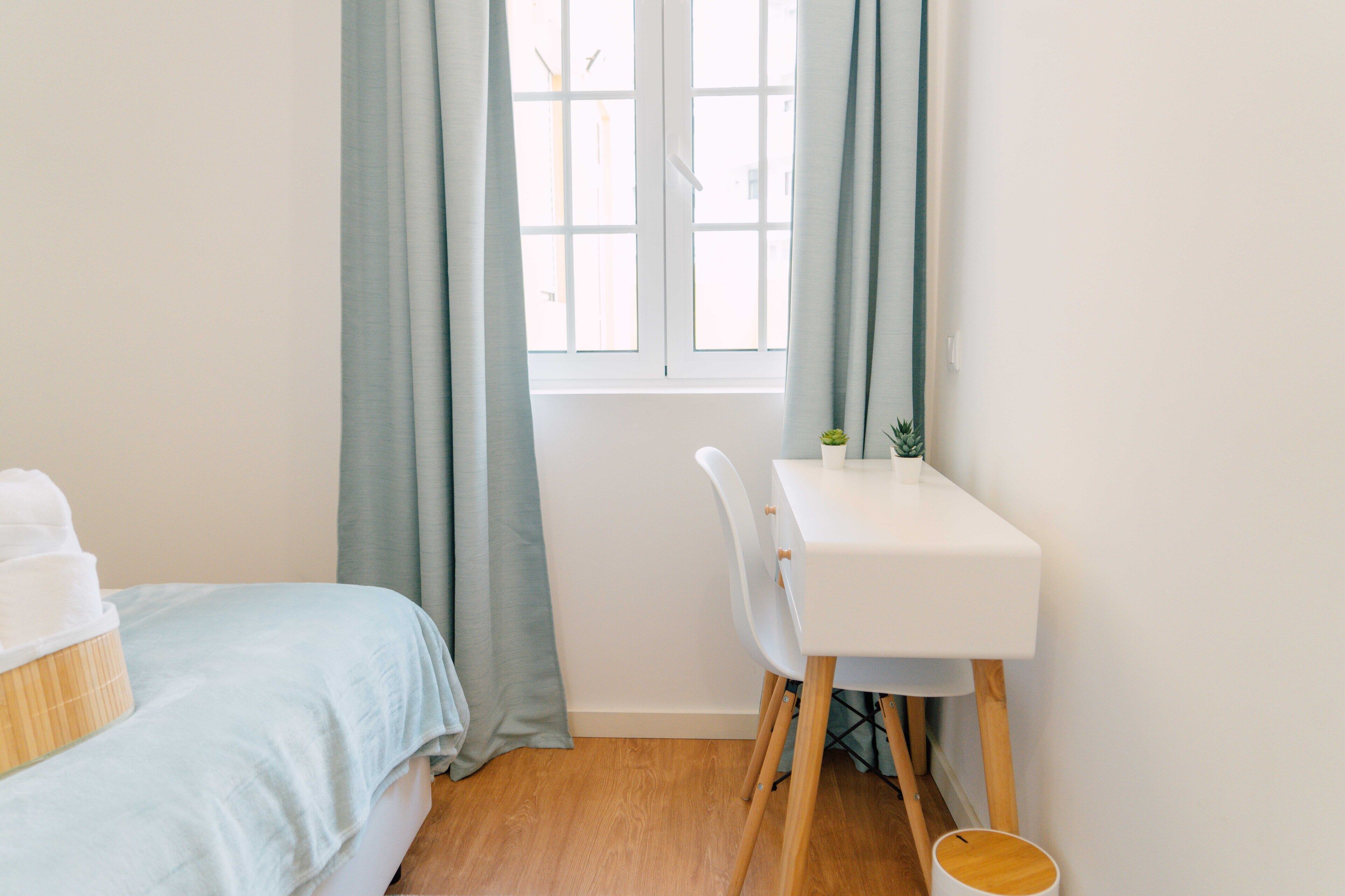 Un dormitorio con cama, un escritorio pequeño con silla y una ventana con cortinas.